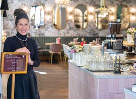 Gastgeberin begrüßt Gäste im stilvollen Café mit Spiegelwand und Blumen