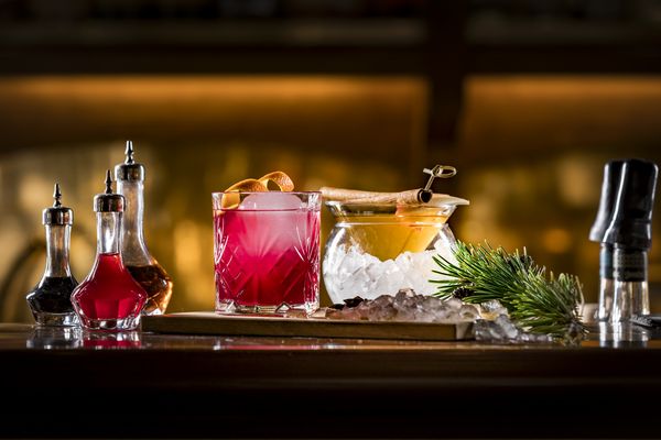 A red cocktail with an orange zest on top and an orange cocktail with cinnamon sticks.