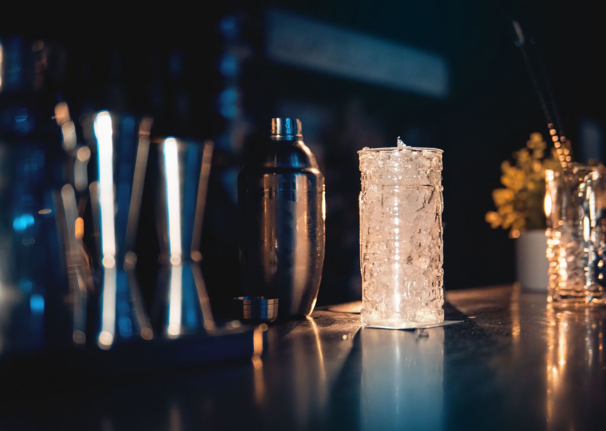 Ein Highball Glas gefüllt mit Eis steht neben einem Shaker in einer Bar.
