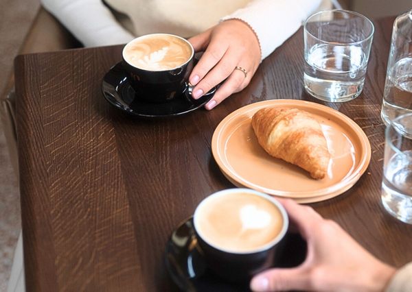 Two cappuccinos, a croissant and glasses of water on a dark brown wooden table.