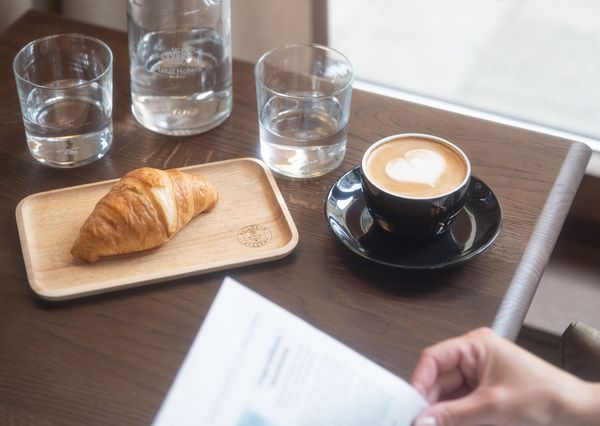 A cappuccino with a heart in the milk foam, a croissant and a few glasses of water are placed on a brown wooden table while the newspaper is read.